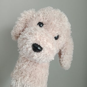 Puede incluir: Un perro de peluche con pelaje marrón claro, ojos negros y nariz negra. El perro lleva un gorro de punto.