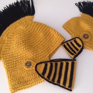May include: Handmade crochet hats in yellow and black, resembling a Roman helmet. The hats feature a black fringe and a decorative button. Two matching striped pieces are also included.