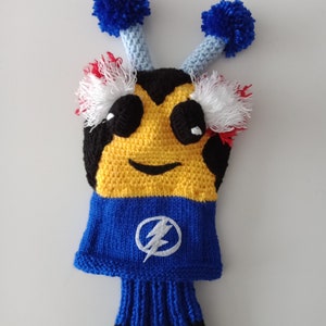 Puede incluir: Un cubre-cabeza de palo de golf de crochet con forma de abeja con pompones azules en la parte superior y un cuerpo azul y amarillo. La abeja tiene un rayo negro en su cuello azul.