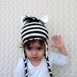 May include: A child wearing a black and white striped zebra hat with long ear flaps and a black mane. The hat is crocheted and has a white and black striped pattern.