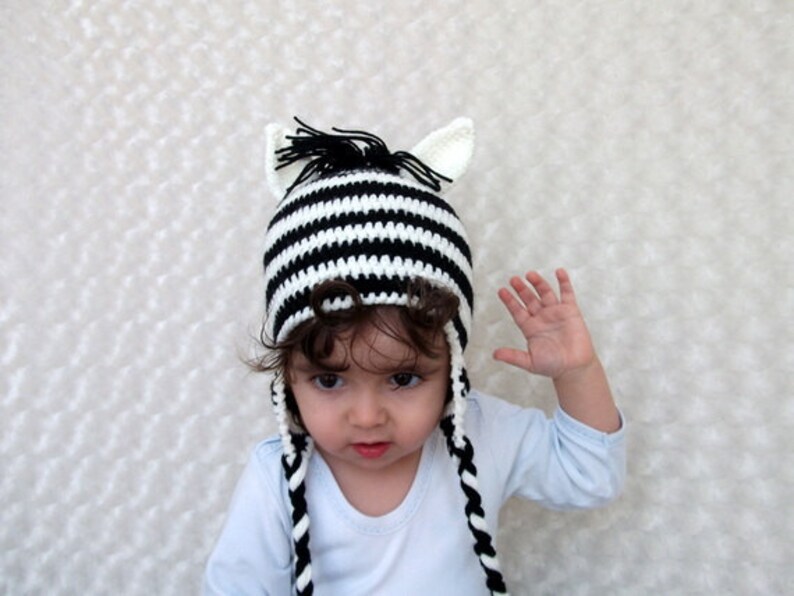 May include: A child wearing a black and white striped zebra hat with long ear flaps and a black mane. The hat is crocheted and has a white and black striped pattern.