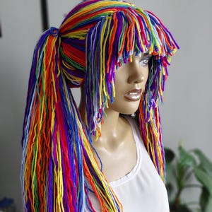 May include: A mannequin head is adorned with a colourful, handmade wig. The wig is made of long strands of yarn in a rainbow of shades, including red, orange, yellow, green, blue, and purple. The wig is styled in pigtails.