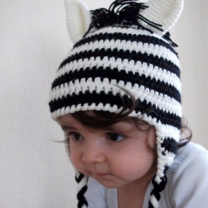 May include: A black and white striped crocheted zebra hat with long ear flaps and a black mane. The hat is worn by a child with brown hair.