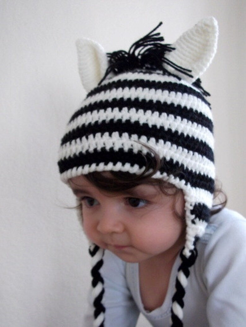 May include: A black and white striped crocheted zebra hat with long ear flaps and a black mane. The hat is worn by a child with brown hair.