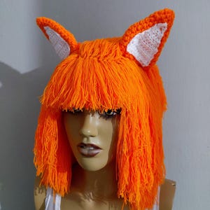 Pode incluir: Um chapéu de raposa de fio laranja com orelhas internas brancas. O chapéu apresenta uma franja de fio laranja e foi projetado para se assemelhar a uma raposa. O chapéu é exibido em uma cabeça de manequim.