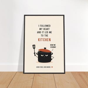 Op de afbeelding: Een ingelijste kunstprint met een crèmekleurige achtergrond met de tekst I FOLLOWED MY HEART AND IT LED ME TO THE KITCHEN. Daaronder een zwarte pan met een oranje deksel die een spatel vasthoudt, met de tekst NOW WE COOKING! en GOOD FOOD, GOOD MOOD.
