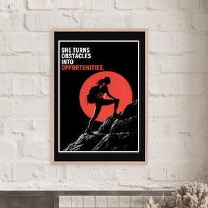 May include: A framed vertical poster featuring a black silhouette of a hiker with a backpack climbing a rocky mountain against a large red sun. The text in white and red reads: SHE TURNS OBSTACLES INTO OPPORTUNITIES. The design is set on a black background.