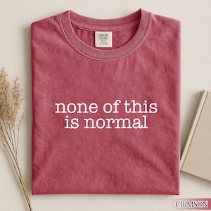 May include: Crimson-colored t-shirt with the phrase "none of this is normal" printed in white. The shirt is folded and has a Comfort Colors tag at the neckline. A book and dried plant stems are in the background.