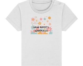 May include: White short-sleeved t-shirt with a colorful graphic. The graphic features flowers, stars, and the text "WANN KOMMT'S SCHOPPERLE?" in a playful font. The shirt has a round neckline and a snap closure on the shoulder.