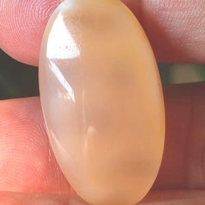 May include: A polished, oval-shaped gemstone with a creamy, peach-colored hue. The stone has a smooth, glossy surface, reflecting light. The gemstone is held between fingers, showcasing its size and shape. This is a close-up of a natural gemstone.