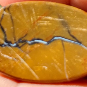 May include: Oval-shaped gemstone with a golden-yellow base color. The stone features a network of dark gray and white veins, with a striking light blue vein running across the center. The surface has a polished appearance.