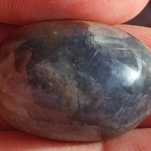 May include: A polished, oval gemstone displaying a blend of colours. The stone features a dark blue base, interwoven with swirls of brown, tan, and white. The surface is smooth, reflective, and has a glossy finish.
