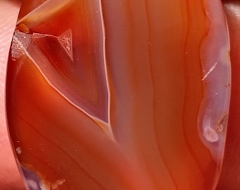 May include: Oval-shaped polished gemstone with a mix of red, orange, and white bands. The stone has a smooth, glossy surface and a natural, organic appearance. The gemstone is a unique piece of jewelry.