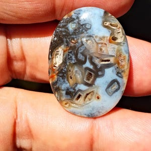 Könnte beinhalten: Ein ovaler Edelstein mit hellblauem Grund, der komplizierte Muster in Braun-, Schwarz- und Goldtönen aufweist. Die Oberfläche des Steins ist poliert und hebt die natürlichen Variationen und Texturen des Materials hervor. Geeignet für Schmuck.