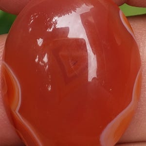 May include: A polished, oval-shaped gemstone in shades of red-orange with white and orange banding. The stone has a smooth, glossy surface and is likely a cabochon, suitable for jewelry making or collecting.
