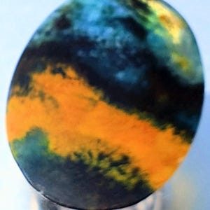 May include: An oval gemstone displaying a vivid mix of colours. The stone showcases a striking blend of blue, yellow, and orange tones, with dark detailing. The polished surface reflects light, emphasising the intricate patterns and colour variations.