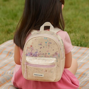Peut inclure: Un sac à dos beige avec une fermeture éclair et des bretelles roses, décoré d'étoiles colorées et de motifs floraux. Le sac à dos est porté par une personne vêtue d'une robe rose, assise sur une couverture à carreaux sur l'herbe.