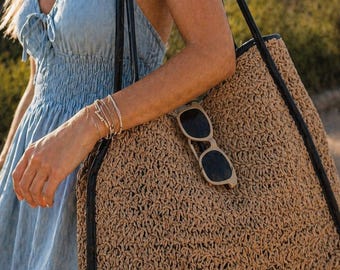 May include: A large, woven tote bag in a natural tan color with black trim and straps. Sunglasses rest on the bag's surface. The bag is carried over the shoulder. The image also shows a light blue dress.