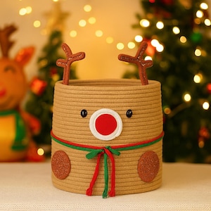 May include: A tan rope basket decorated as a reindeer with brown antlers, black eyes, and a red nose. The basket has a green and red ribbon around the middle and brown circular patches on the sides. Festive holiday decor.