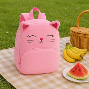 Może przedstawiać: Różowy pluszowy plecak w kształcie kota z kieszenią zapinaną na zamek błyskawiczny i haftowanym wizerunkiem kota.