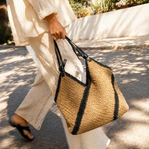 Peut inclure: Un grand sac cabas en paille tressée beige, orné de larges bandes verticales noires et doté de bretelles en cuir noir nouées aux points d'attache supérieurs. Le sac présente une forme structurée et angulaire avec une large ouverture.