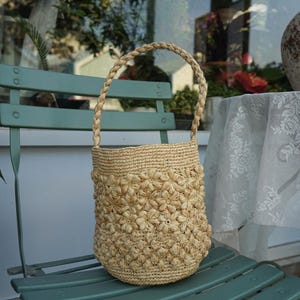 May include: A natural-toned woven straw handbag with a braided handle. The body of the bag is adorned with a floral pattern. The bag is sitting on a teal green chair.
