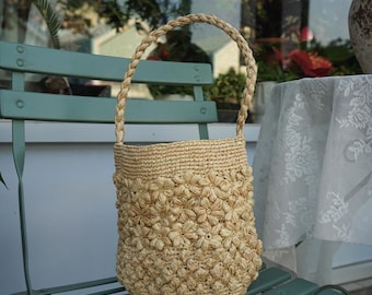 May include: A natural-toned woven straw handbag with a braided handle. The body of the bag is adorned with a floral pattern. The bag is sitting on a teal green chair.