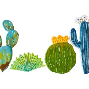 May include: A set of five ceramic cactus wall decor pieces. The set includes a tall blue cactus with white flowers, a round green cactus with a yellow crown, a green fan-shaped cactus, and two blue and green prickly pear cactus pieces.
