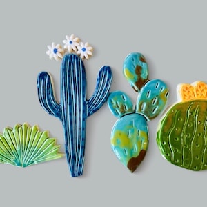 May include: Five ceramic cactus tiles in various shapes and colors. A blue cactus with white flowers, a green cactus with yellow accents, and three green and blue prickly pear cactus tiles.