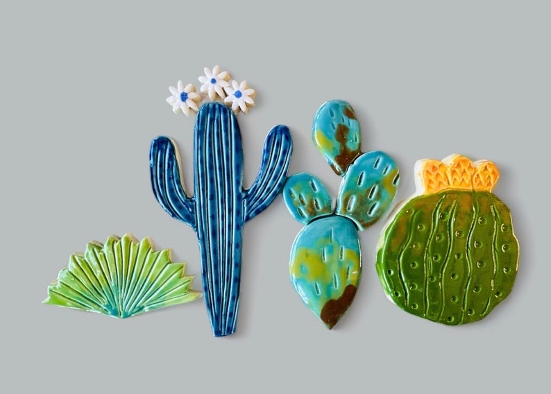 May include: Five ceramic cactus tiles in various shapes and colors. A blue cactus with white flowers, a green cactus with yellow accents, and three green and blue prickly pear cactus tiles.