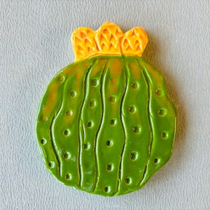 May include: A green ceramic cactus with a yellow flower on top. The cactus has a textured surface with small bumps and lines.