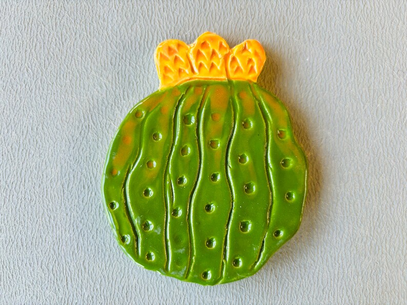 May include: A green ceramic cactus with a yellow flower on top. The cactus has a textured surface with small bumps and lines.