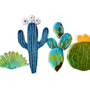 May include: A set of five ceramic cactus tiles in various shades of green, blue, and yellow. The tiles feature different cactus shapes, including a tall saguaro cactus with white flowers, a prickly pear cactus, and a round barrel cactus.