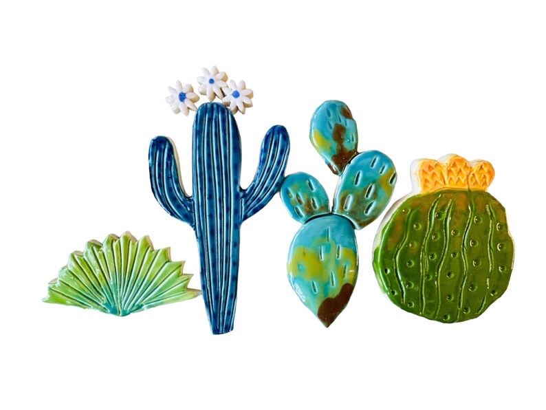 May include: A set of five ceramic cactus tiles in various shades of green, blue, and yellow. The tiles feature different cactus shapes, including a tall saguaro cactus with white flowers, a prickly pear cactus, and a round barrel cactus.