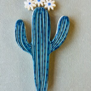 May include: A blue ceramic cactus with white flowers on top. The cactus has a textured surface with lines and dots.