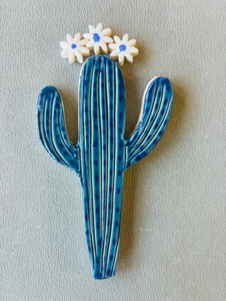 May include: A blue ceramic cactus with white flowers on top. The cactus has a textured surface with lines and dots.