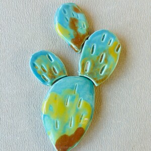 May include: A ceramic cactus sculpture with three sections. The cactus is painted in shades of blue, green, yellow, and brown.