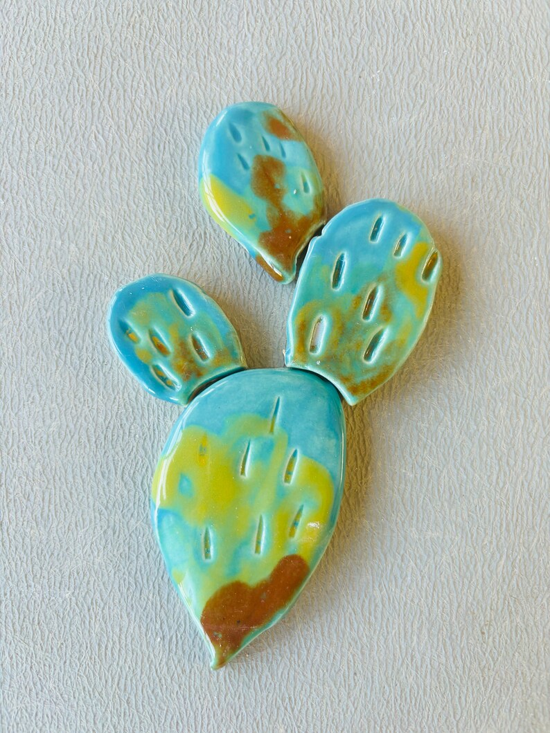 May include: A ceramic cactus sculpture with three sections. The cactus is painted in shades of blue, green, yellow, and brown.
