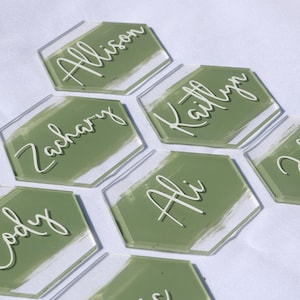 May include: Clear acrylic hexagon place cards with names in white script. Each card has a painted olive green background. Names include Allison, Kaitlyn, John, Ali, Cody, and Zachary. Ideal for weddings or events.