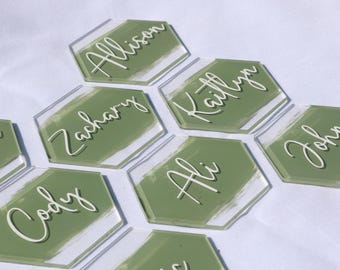 May include: Clear acrylic hexagon place cards with names in white script. Each card has a painted olive green background. Names include Allison, Kaitlyn, John, Ali, Cody, and Zachary. Ideal for weddings or events.