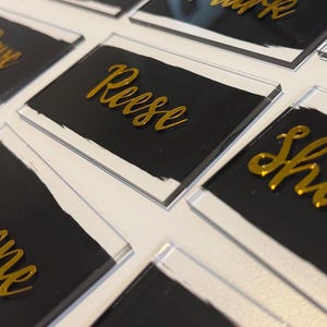 May include: Close-up of several name cards. Each card has a clear acrylic base with a black painted section and a gold-colored name in a cursive font. The names are different on each card.