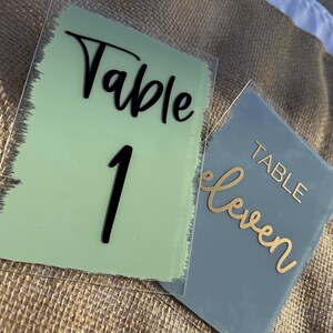 May include: Two acrylic table number signs. One is light green with black text that reads "Table 1". The other is gray with gold text that reads "Table Eleven". Both signs have a clear border.