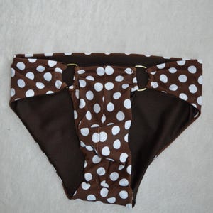 May include: A brown bikini bottom featuring a white polka dot pattern throughout. The garment includes two gold-toned metal rings connecting the front and side panels, with a solid brown interior lining visible along the leg openings and waistband.