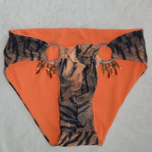 May include: Bikini bottoms in a vibrant orange hue featuring a dark brown tiger stripe pattern on the side panels and center. Two silver-toned metal rings connect the sections, each adorned with dangling amber-colored beads and small charms.