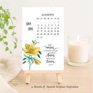 Op de afbeelding: Een kleine, witte kalender voor augustus, weergegeven op een houten ezel. De kalender heeft een gele bloemenillustratie en Spaanse tekst, inclusief een bijbelvers. Een brandende kaars staat rechts. De tekst luidt "12 Months of Spanish Scripture Inspiration".
