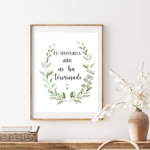 May include: A framed print with the text "TU HISTORIA aún no ha terminado;" in black script, surrounded by a green leafy wreath. The print is in a wooden frame and is displayed on a wooden surface.