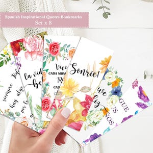 May include: Set of eight Spanish inspirational quote bookmarks. Each bookmark features floral watercolor designs with phrases like "Vive cada momento" and "Sonríe!" in elegant script. The bookmarks are held in a hand.