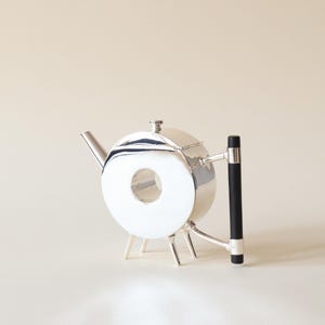 Könnte beinhalten: Eine silberne Teekanne mit einem runden Körper und einem schwarzen Griff. Die Teekanne hat ein einzigartiges Design mit vier Beinen und einem Loch in der Mitte des Körpers.