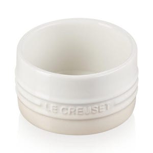 May include: A white and cream-colored ceramic ramekin with the embossed text "LE CREUSET" on the base. The ramekin has a rounded shape with a smooth, glossy finish. It is designed for baking and serving individual portions.