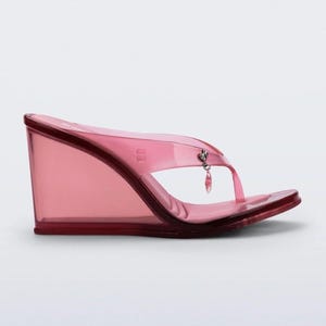 Könnte beinhalten: Transparente, rosa Keilsandalen mit Zehensteg-Design. Die Sandalen haben einen Keilabsatz und einen kleinen Anhänger am Riemen. Die Schuhe sind aus einem transparenten, glänzenden Material.